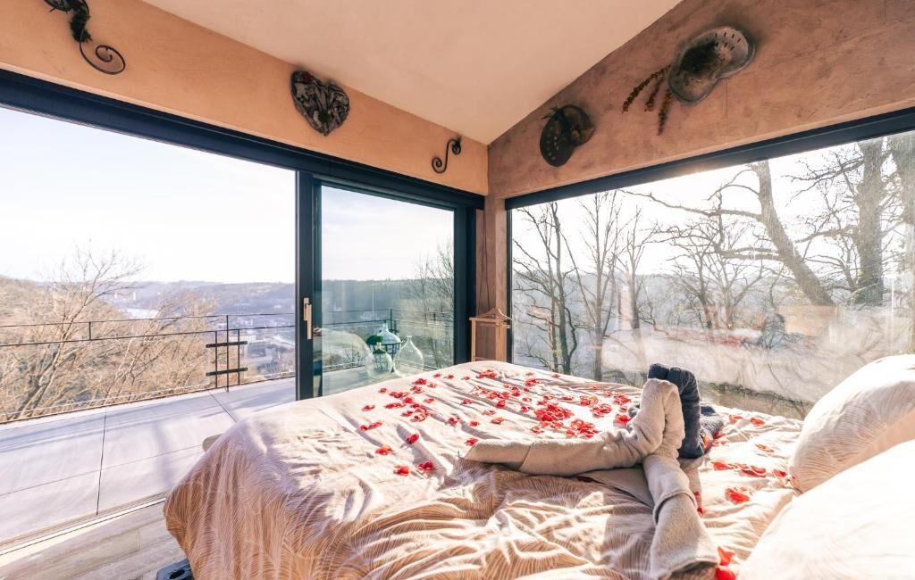 Lit king size avec pétales de roses face à une grande baie vitrée offrant une vue panoramique sur la nature et la vallée de Dinant